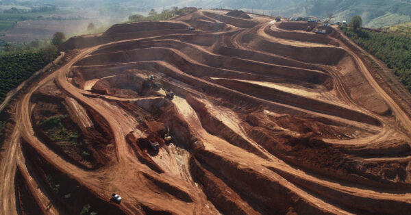 Vista aérea de uma mineração a céu aberto com terra vermelha, mostrando estradas e máquinas de escavação em operação, cercada por vegetação e colinas ao fundo. Fonte: Cabo Verde Mineração.