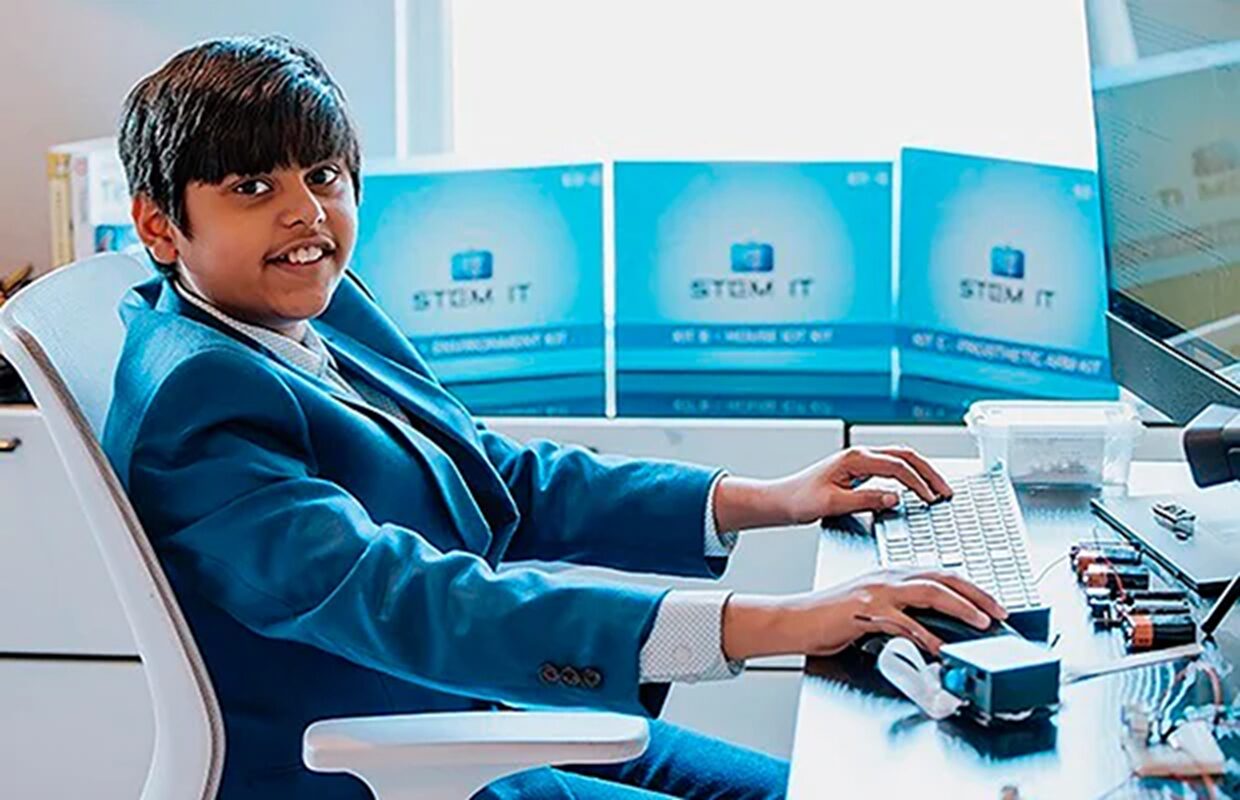 doenças cardíacas - Jovem de 14 anos, usando terno azul, sentado em frente ao computador com kits de eletrônica e caixas de projeto educacional ao fundo.