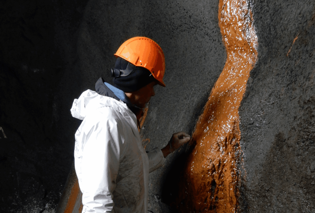 Engenheiro com capacete laranja inspeciona túnel rodoviário onde há sinais de degradação no concreto, com fluxo de líquido enferrujado escorrendo pela parede.