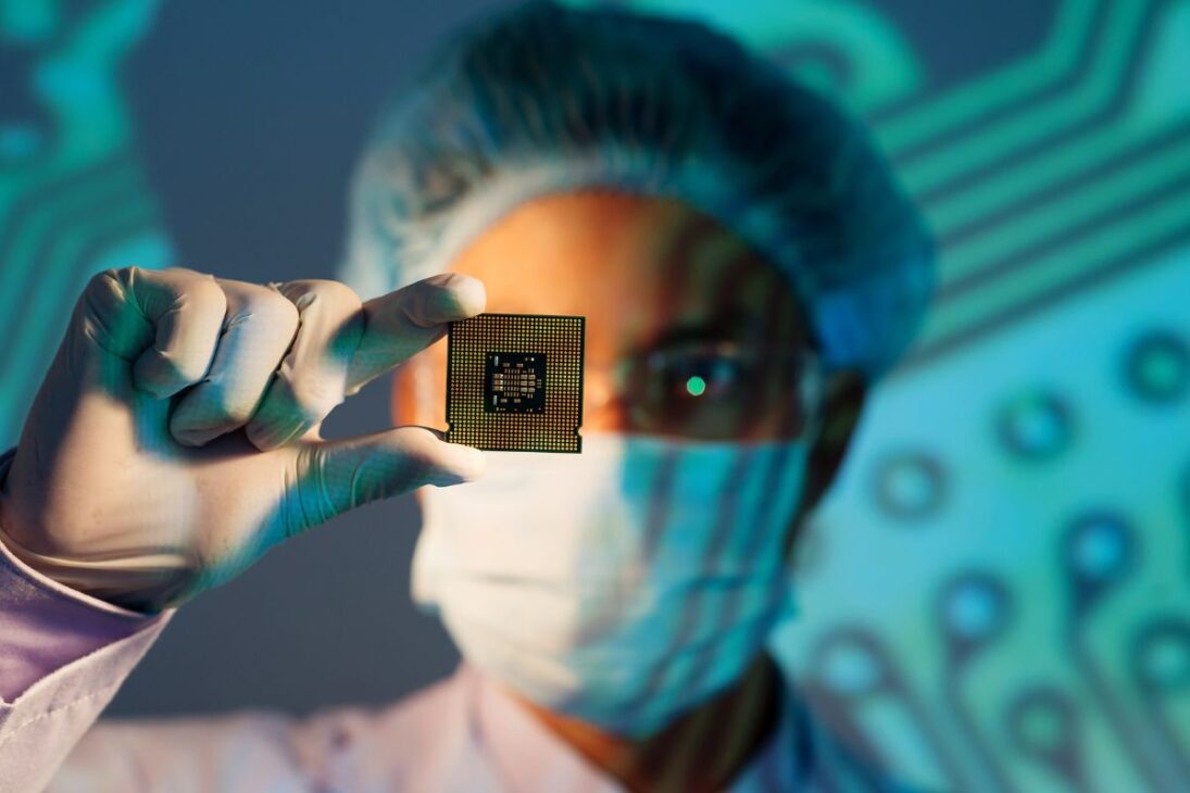 Científico con máscara, gorro y guantes sosteniendo un microchip, representando investigación, innovación y alta tecnología en electrónica.