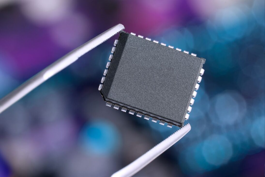 Microchip electrónico siendo cuidadosamente sostenido por unas pinzas, con fondo desenfocado en tonos azulados, representando precisión en la fabricación de componentes de alta tecnología.