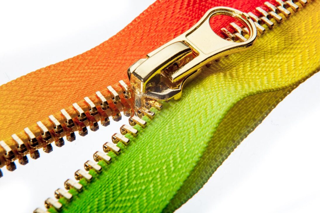 Zíper de metal dorado sobre un fondo blanco, conectando tejidos con colores vibrantes: naranja, verde-limón y amarillo, mostrando la funcionalidad del cierre.