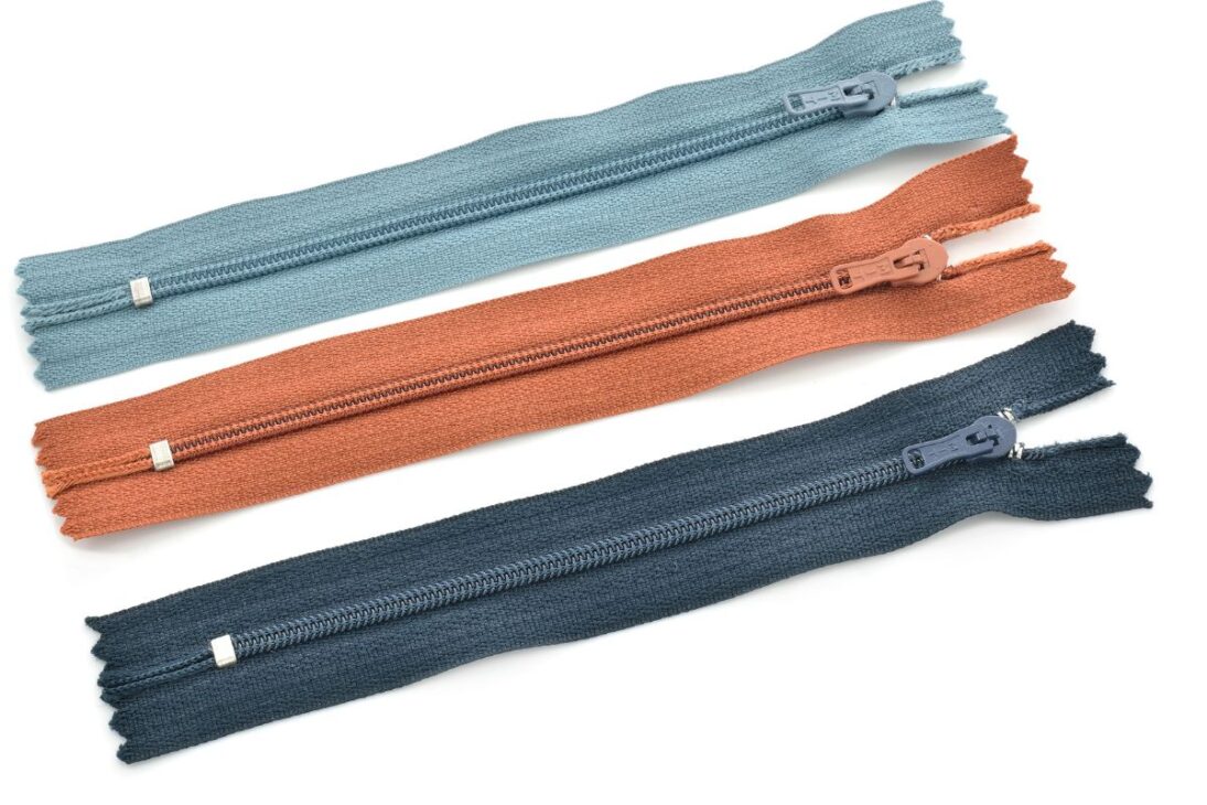 Tres zíperes de nailon coloridos (azul claro, terracota y azul marino), cada uno con su respectivo tirador, dispuestos diagonalmente sobre un fondo blanco.