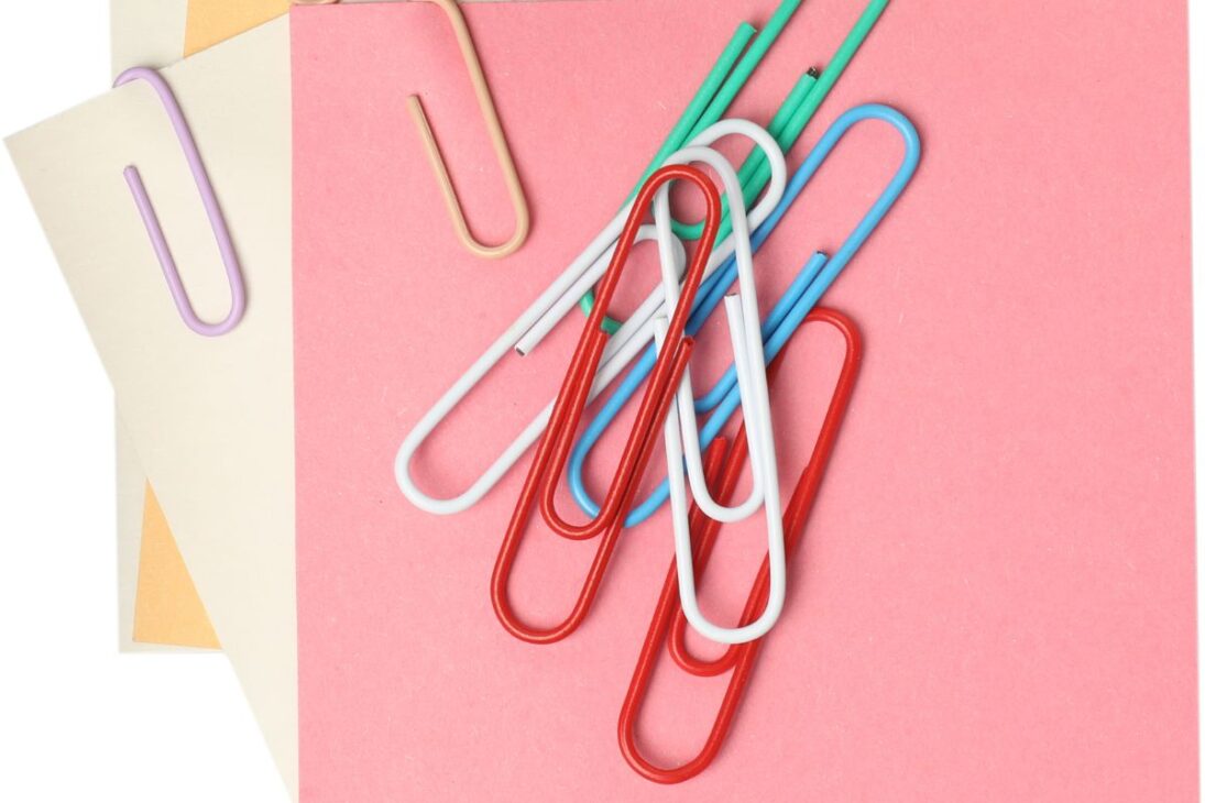 Una mezcla de clips de papel grandes en rojo, blanco y azul, junto con otros colores, sobre papeles rosa y beige.