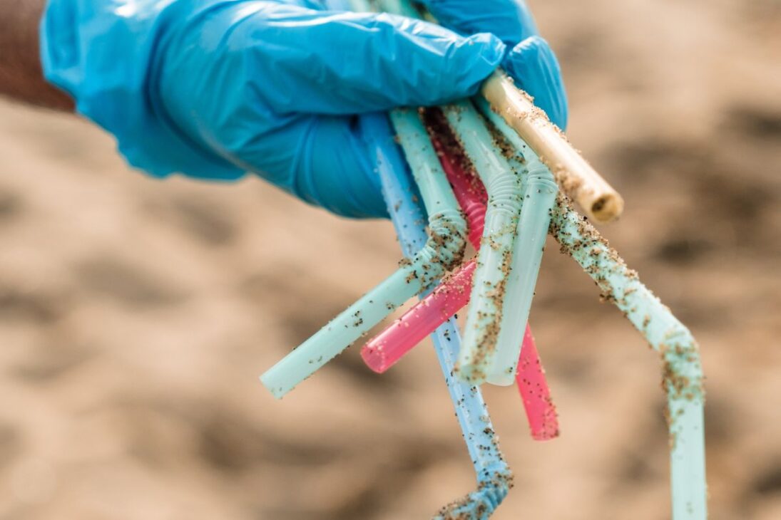 Una mano con guante azul de goma sostiene un puñado de popotes de plástico sucios de arena.