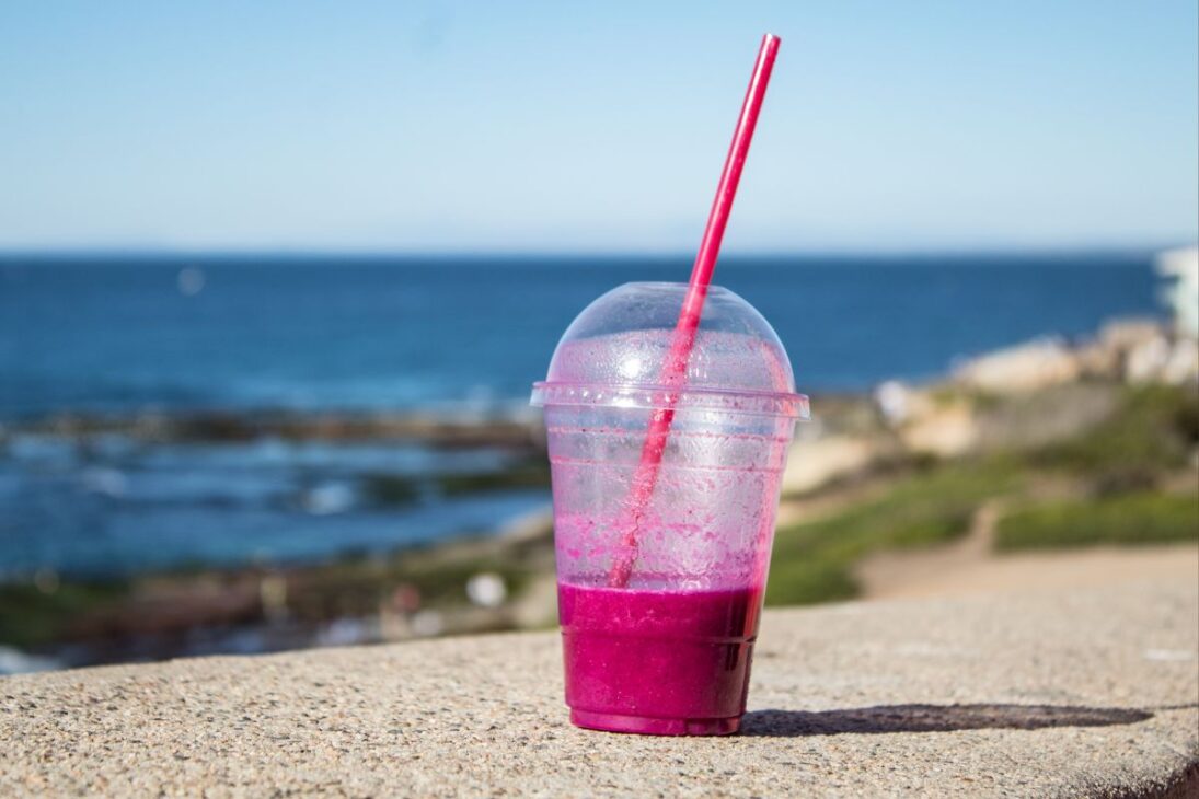 Un vaso de plástico casi vacío con restos de bebida morada y un popote rosa, en un muro con el océano al fondo.