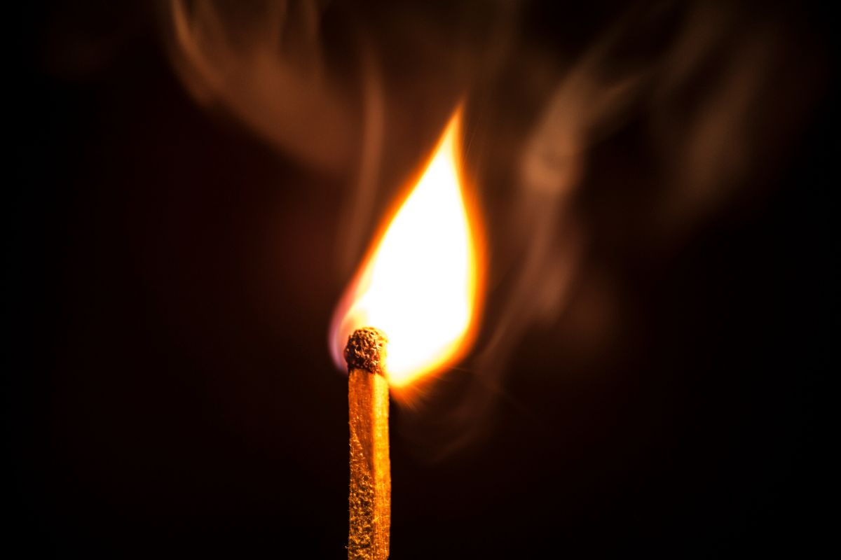 Um único fósforo de madeira com uma chama brilhante e fumaça ondulante contra um fundo preto.