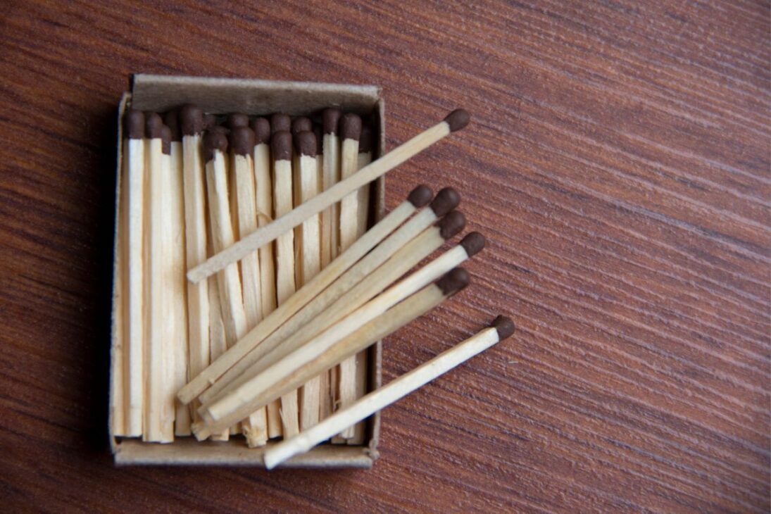 Una caja de fósforos de cartón abierta con varios fósforos de madera dentro y algunos esparcidos sobre una superficie de madera marrón.