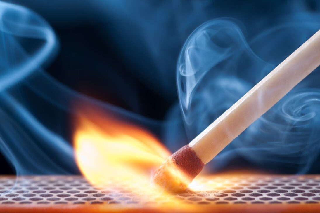 Un fósforo de madera está encendido, con una llama naranja y amarilla vibrante y humo azul ondeando contra un fondo oscuro y una caja de fósforos texturizada.