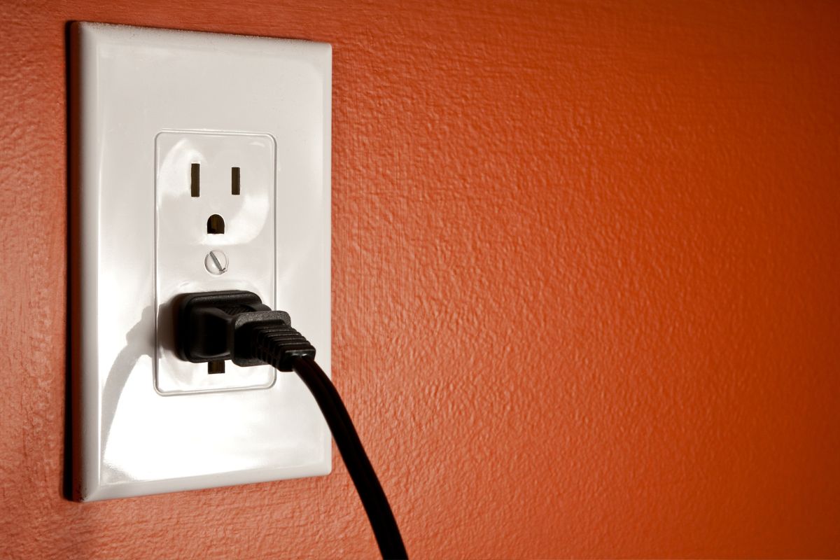 Plugue preto inserido na parte inferior de uma tomada elétrica branca em uma parede laranja.
