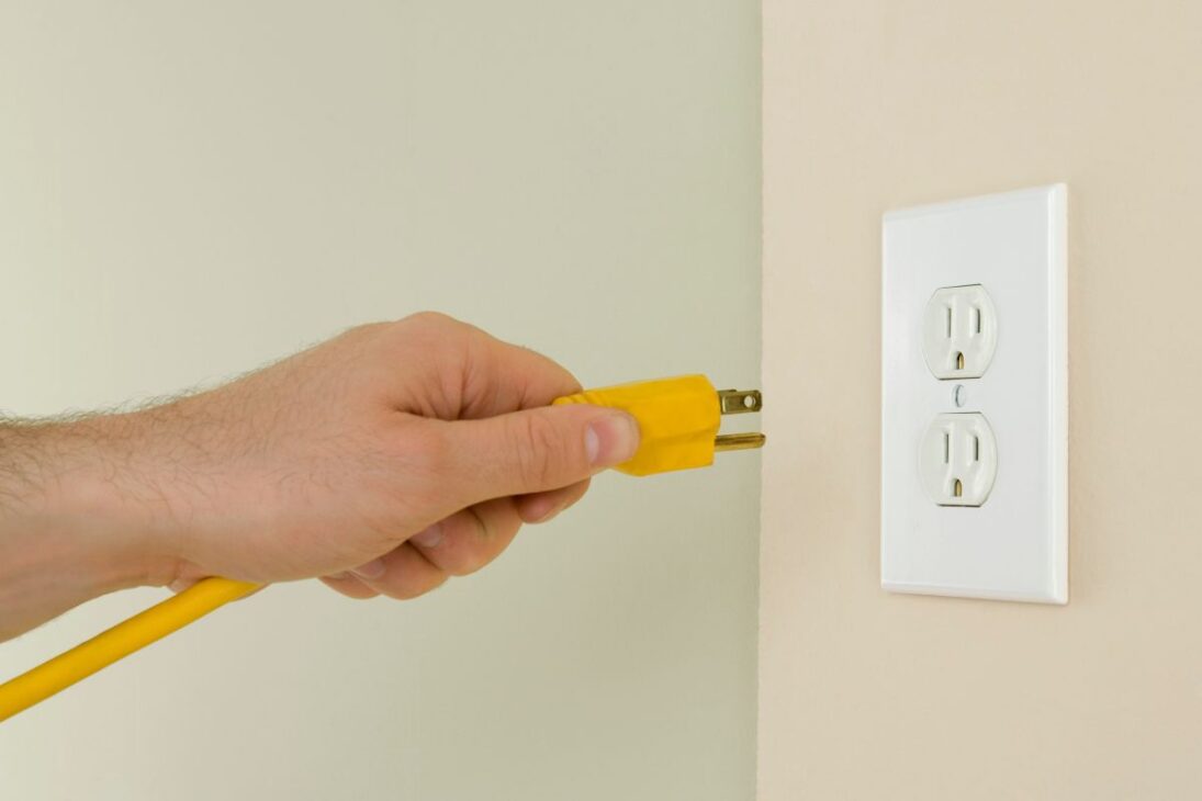 Mano insertando un enchufe amarillo de tres pines (Tipo B) en una toma eléctrica americana blanca de dos sockets.