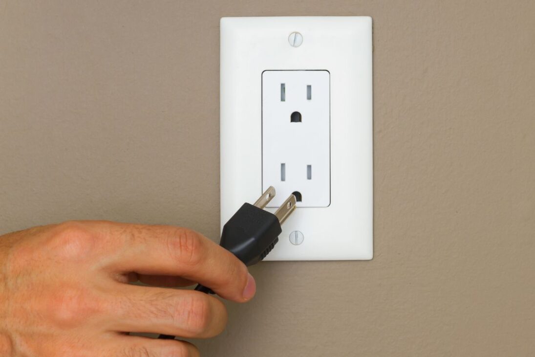 Mano sosteniendo un enchufe negro de dos pines (Tipo A) a punto de ser insertado en una toma eléctrica blanca de dos sockets.