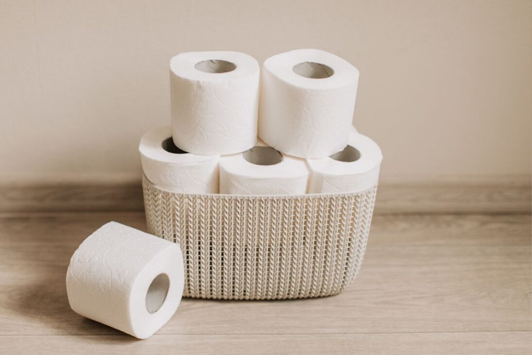 Cesto de mimbre con rollos de papel higiénico blanco apilados en un piso de madera clara.