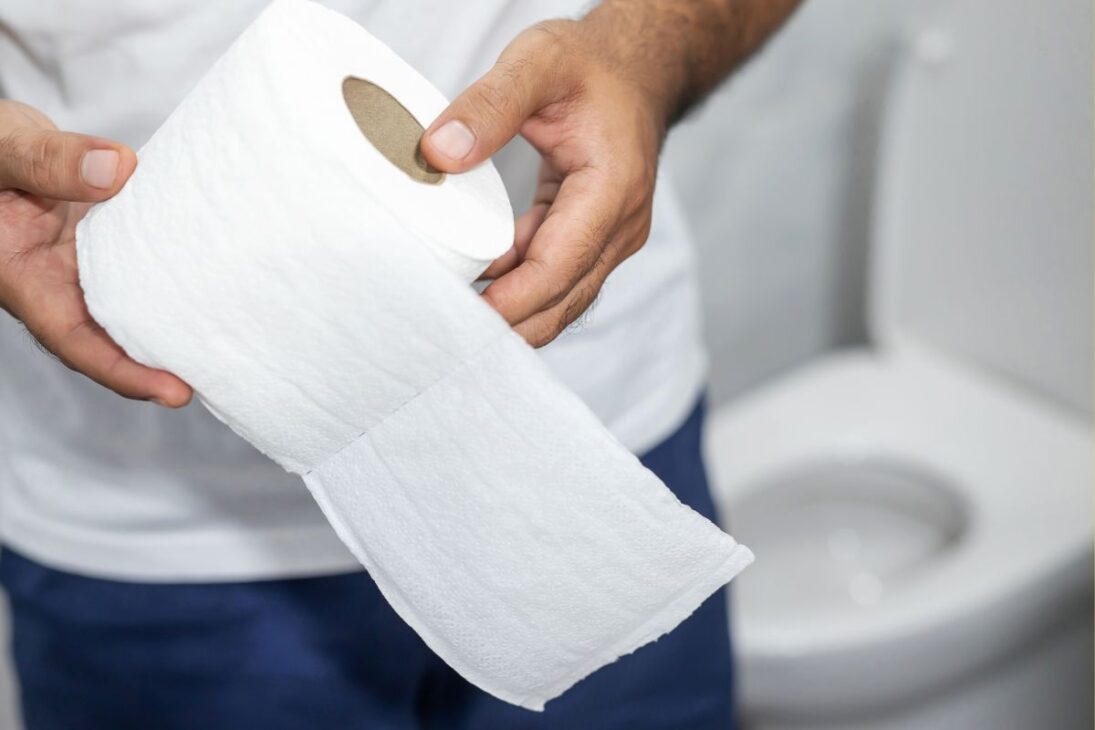 Manos sosteniendo un rollo de papel higiénico con un inodoro desenfocado al fondo.