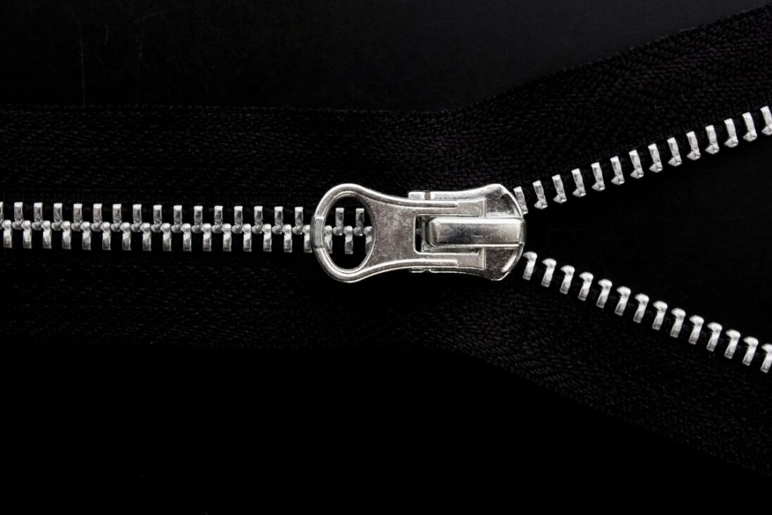 Cierre plateado abierto en una tela negra, mostrando los dientes metálicos brillantes y el tirador en una composición horizontal.