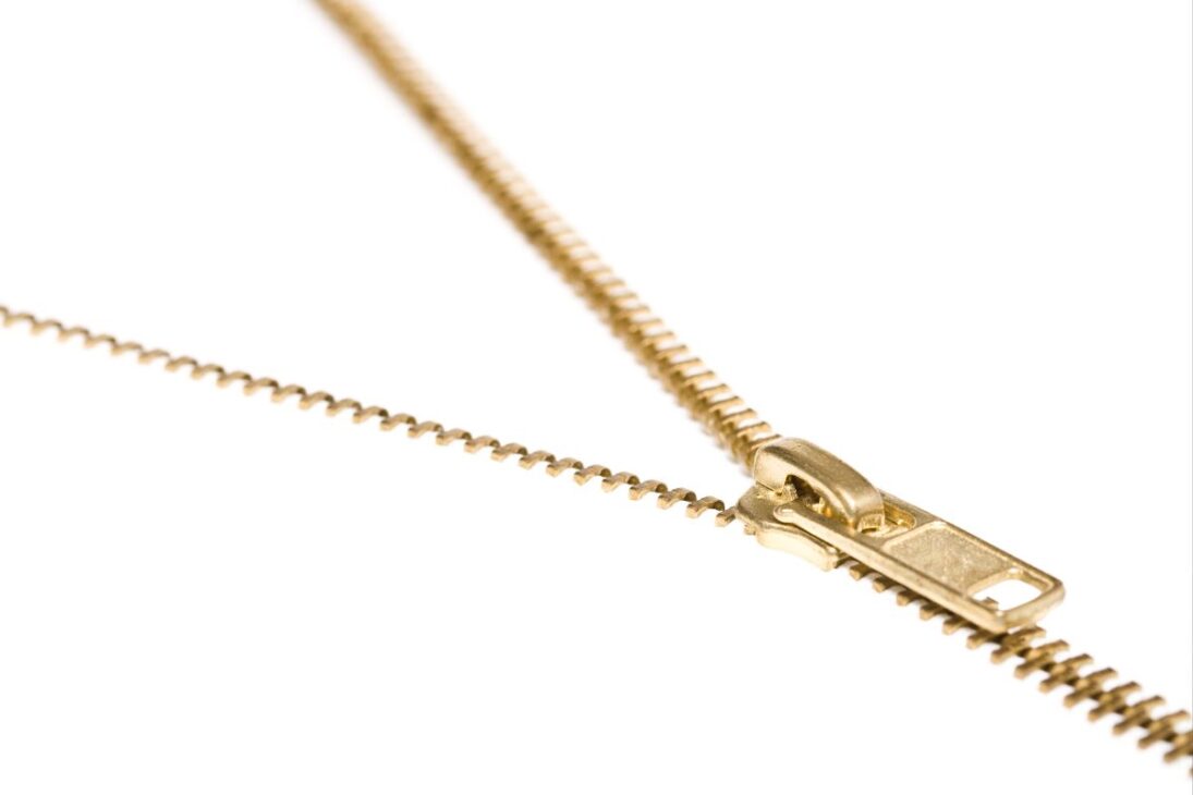 Cierre dorado abierto sobre fondo blanco, con la parte inferior desenganchada y la parte superior unida, creando una composición diagonal.