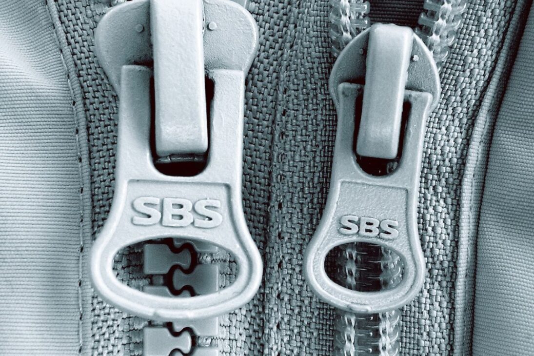  Dos tiradores de zipper grises, marcados "SBS", en tamaños ligeramente diferentes, sobre un tejido texturizado de color claro.