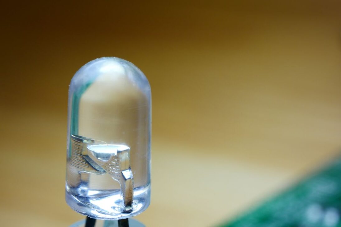 Un diodo emisor de luz (LED) transparente con enfoque en los componentes internos, como el chip semiconductor y el terminal de plomo, contra un fondo desenfocado de madera y circuito verde.