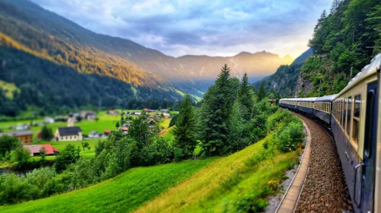 Tren de transporte ferroviario de lujo serpenteando por los Alpes europeos, rodeado de montañas, pueblos y bosques verdes