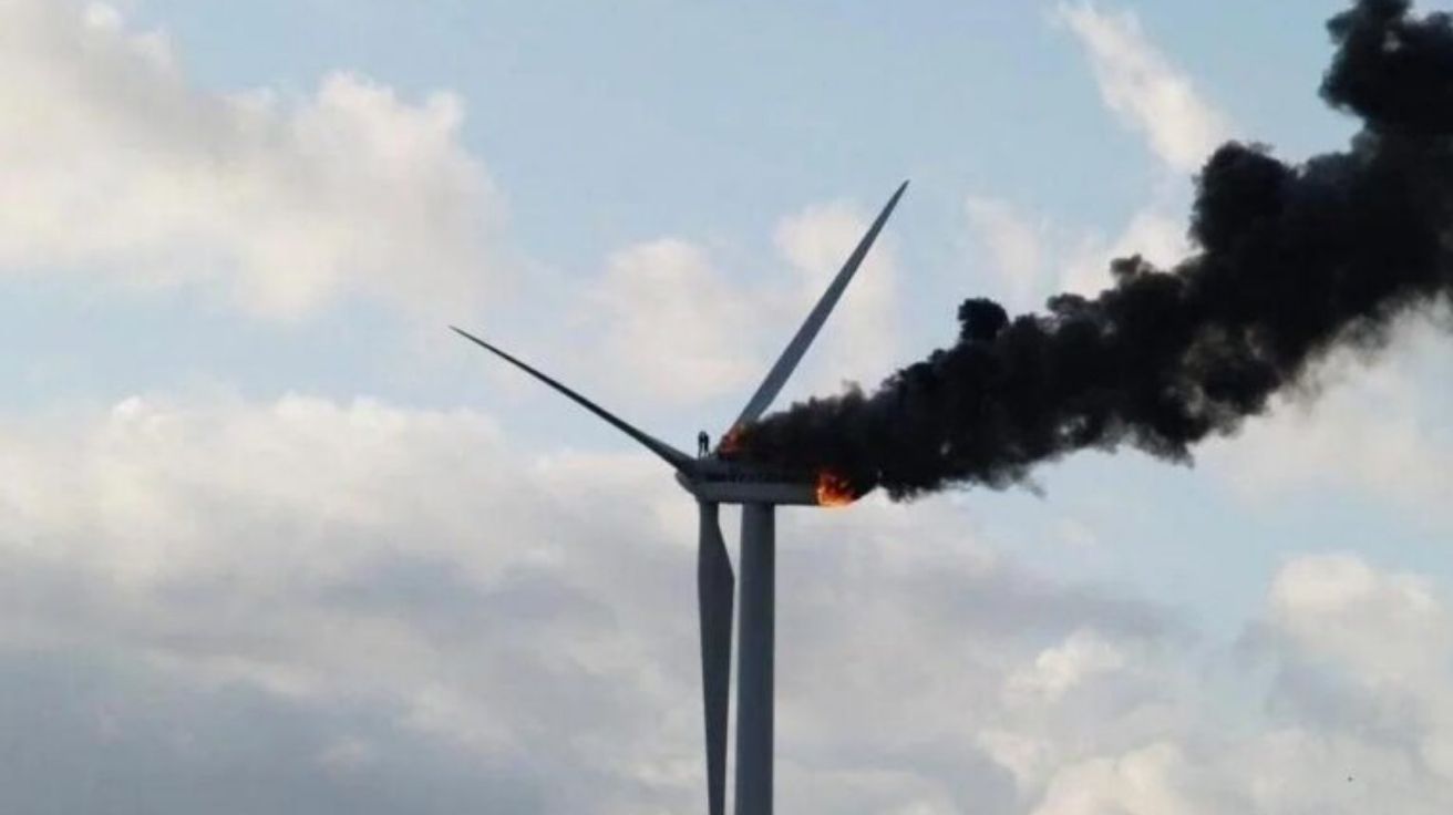 Dois técnicos abraçados no topo de turbina eólica pegando fogo, com fumaça e chamas visíveis.