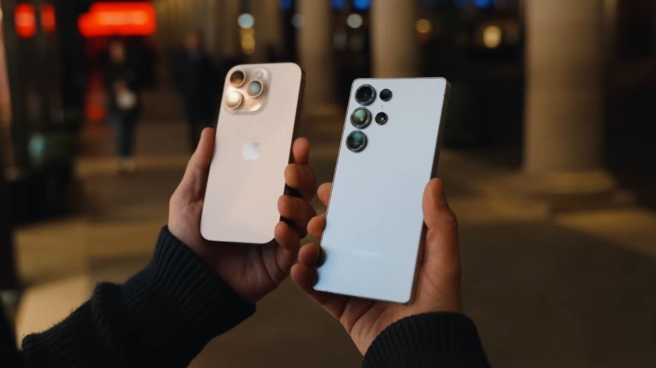 Foto mostrando dois celulares premium, o Galaxy S25 Ultra e o iPhone 16 Pro Max, sendo segurados lado a lado em uma rua iluminada à noite, evidenciando as câmeras e acabamentos.