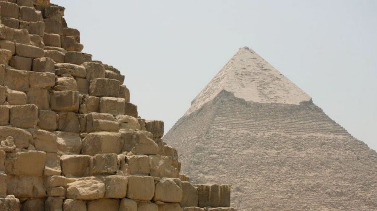 Fotografía real capturada en el complejo de Guiza, en Egipto, muestra en primer plano los enormes bloques de caliza de la Pirámide de Micerinos. Al fondo, destaca la imponente Gran Pirámide de Keops, con parte de su revestimiento original aún visible en la cima. La imagen evidencia la escala monumental y la precisión de la ingeniería egipcia antigua.