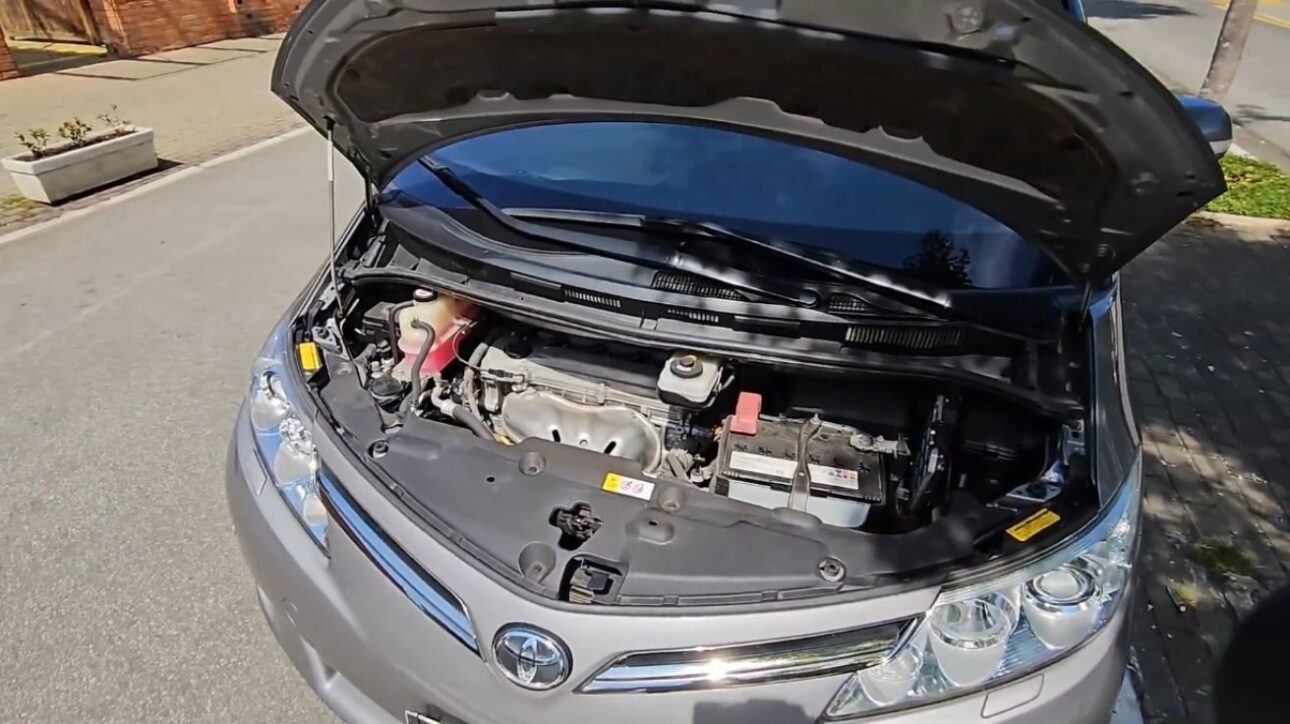 Compartimento del motor de la Toyota Previa 2018 con el capó abierto, exhibiendo el motor 2.4 a gasolina y componentes mecánicos, en una calle urbana durante el día.