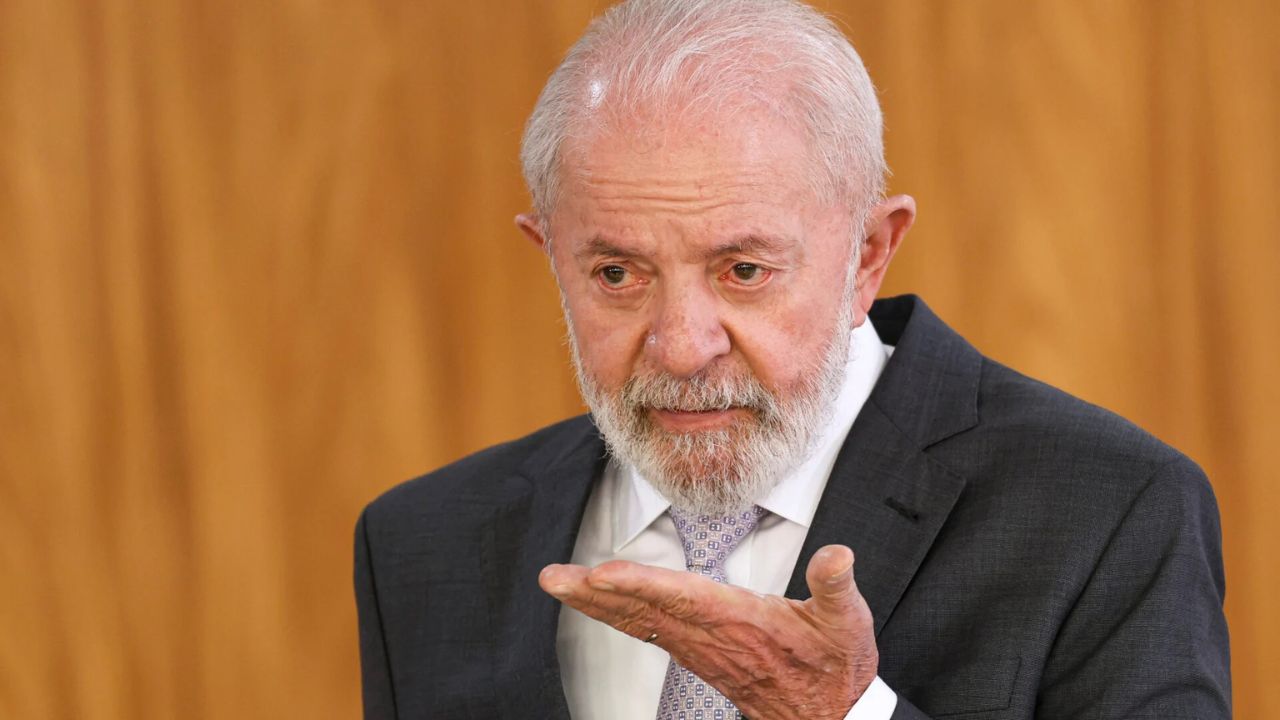 Presidente lula vestindo terno escuro e gravata clara, gesticulando com a mão direita enquanto fala em ambiente formal com fundo de madeira.