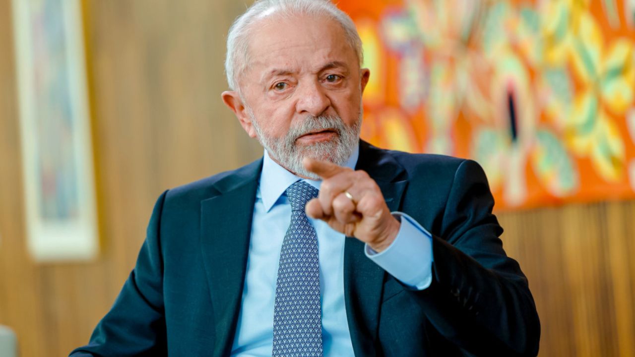 Presidente Lula de terno escuro e gravata, sentado e gesticulando com a mão direita em ambiente formal, com pintura abstrata colorida ao fundo.