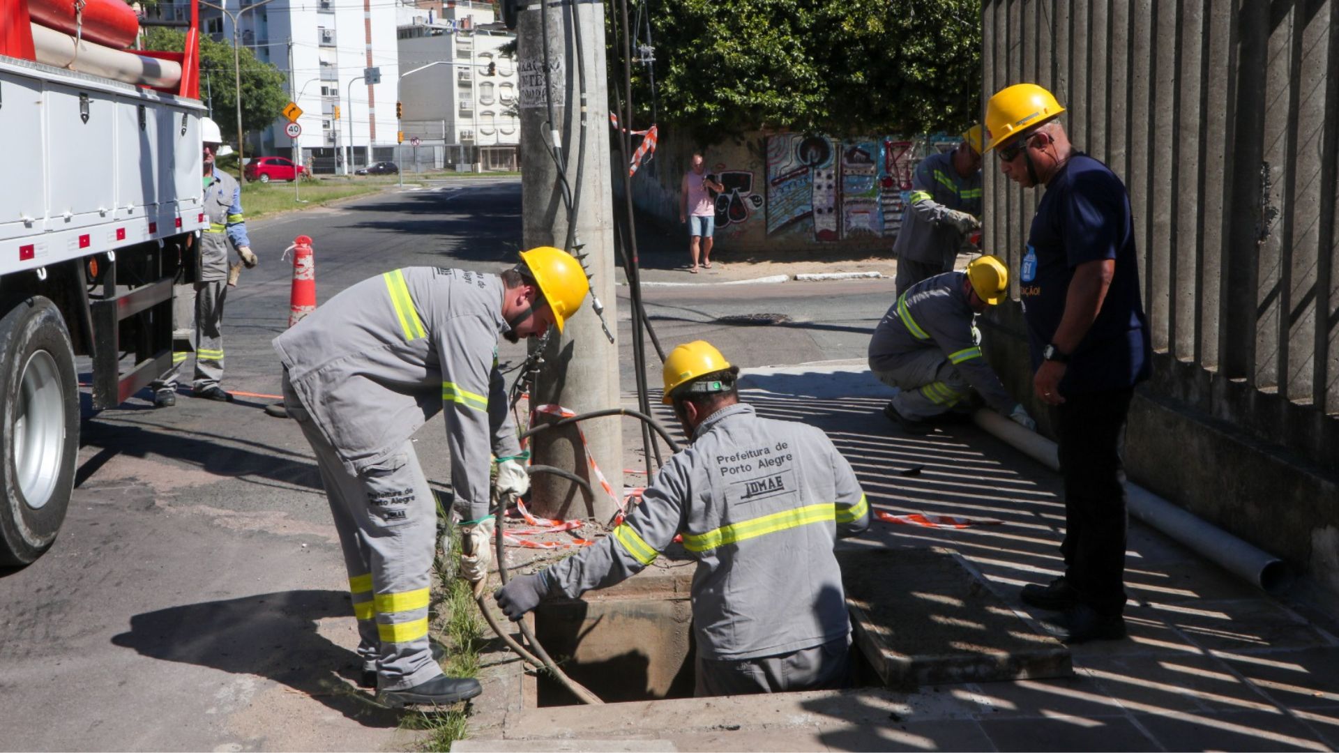 Funcionários do DMAE de Porto Alegre realizam manutenção urbana em um bueiro, utilizando equipamentos de segurança e operando ao lado de um caminhão de serviço em via pública.