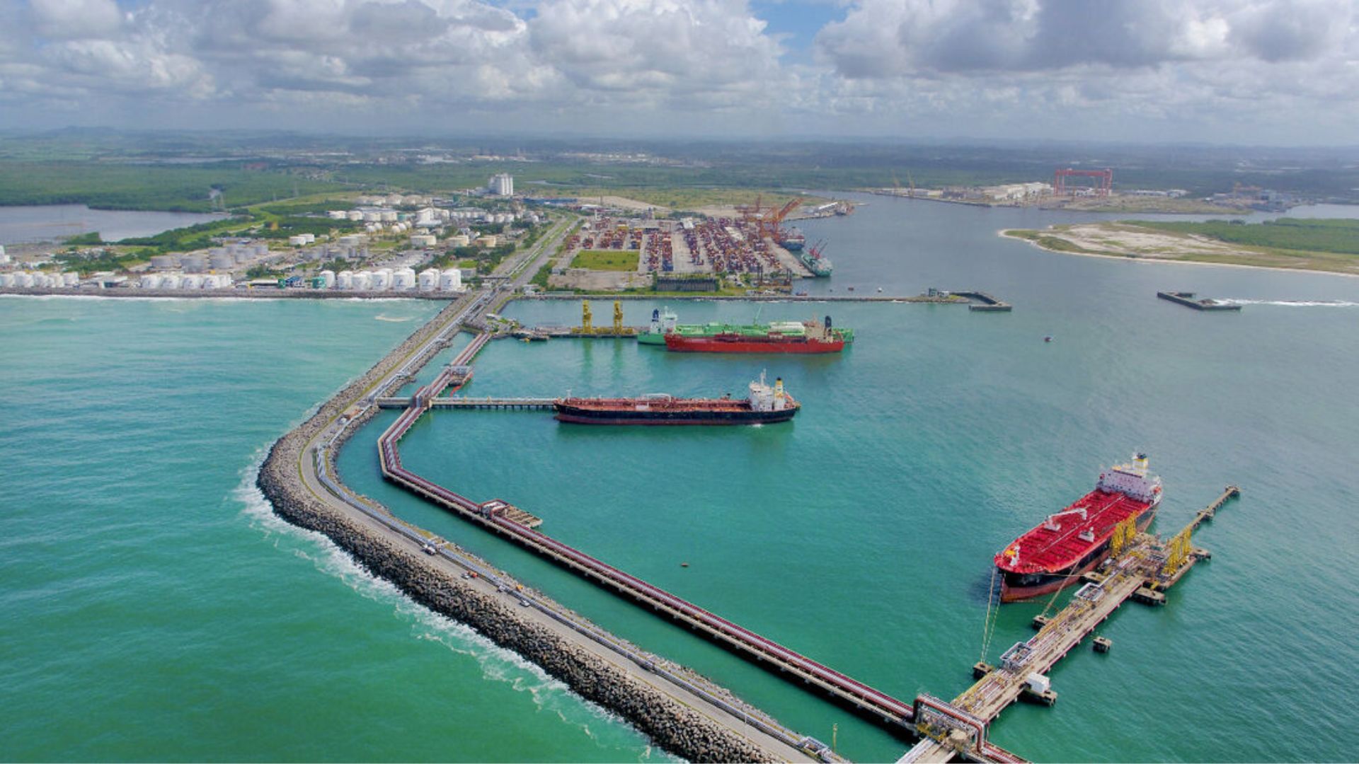 Aérea de terminal portuário com navios cargueiros atracados, tanques de armazenamento e infraestrutura industrial à beira-mar.