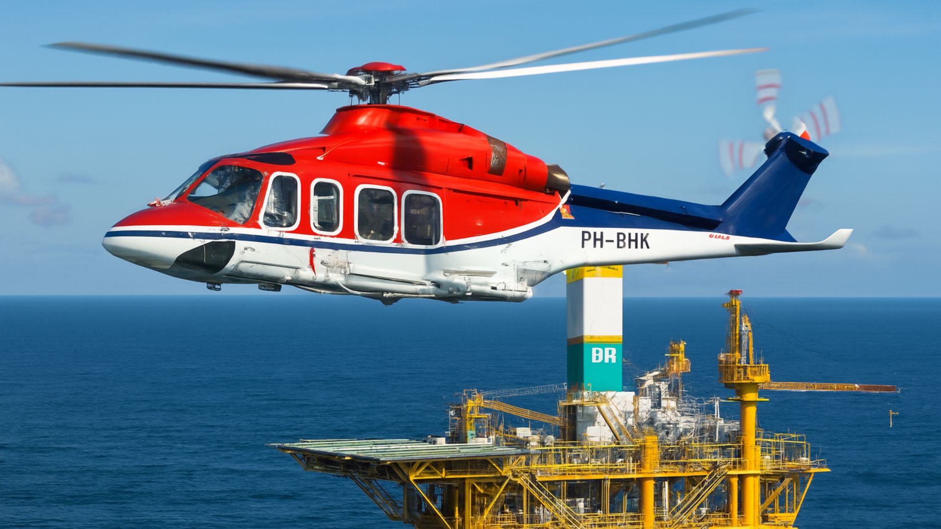 Helicóptero AW139 vermelho e branco em voo sobre uma plataforma de petróleo da Petrobras no oceano, com céu parcialmente nublado ao fundo.