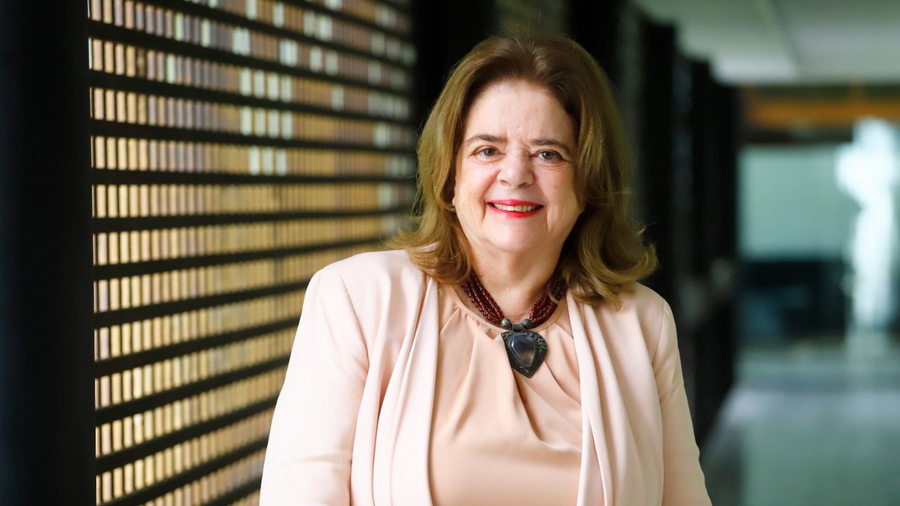 Mulher sorridente em blazer rosa claro, em pé em corredor com parede decorada e piso de azulejos, transmitindo profissionalismo e acolhimento.