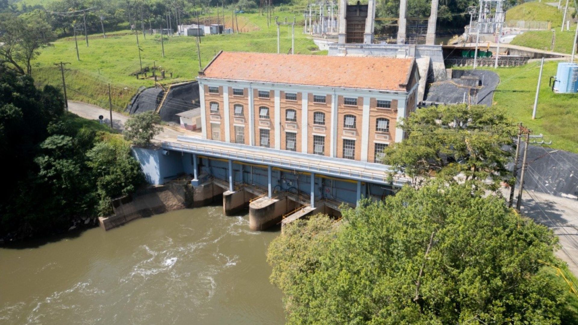 Hidrelétrica com estrutura de alvenaria e comportas sobre o rio, cercada por vegetação, representando geração de energia renovável em área rural.