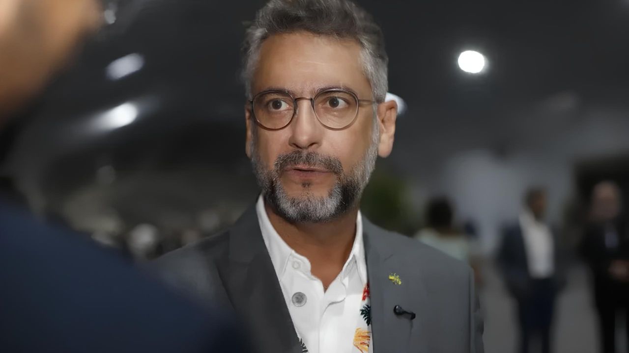 Homem de óculos em traje formal sendo entrevistado em evento profissional, com fundo desfocado.