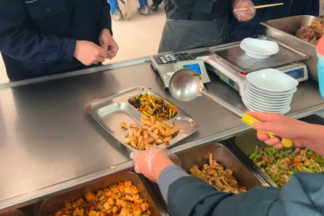 En el comedor de la fábrica china, bandejas de metal sirven almuerzo caliente con fideos al vapor, pollo Kung Pao y rutina intensa de producción para cientos de trabajadores.