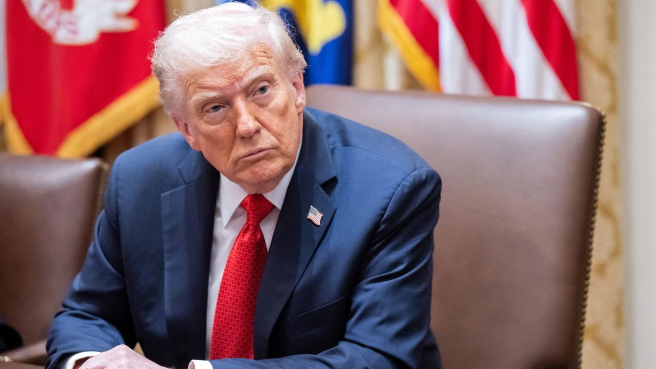 Donald Trump em traje formal sentado à mesa de conferência, com bandeiras dos Estados Unidos ao fundo, em ambiente oficial