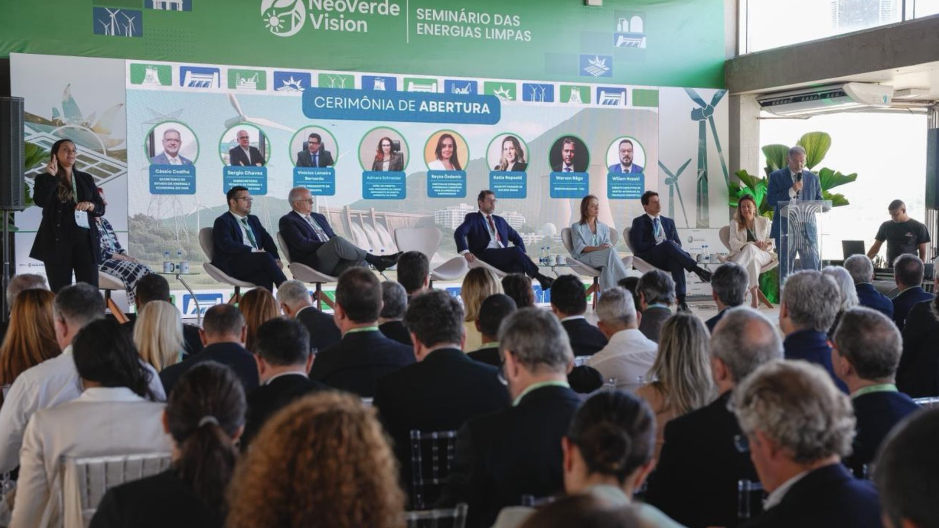 Painel de abertura de um seminário sobre energias limpas, com palestrantes sentados no palco e público assistindo em um auditório iluminado.