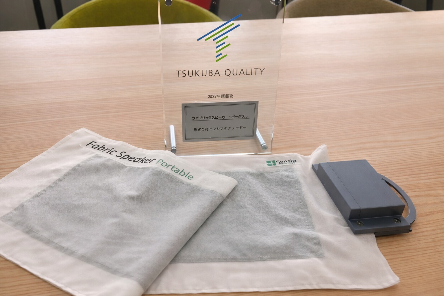 Protótipo de alto-falante em tecido eletrônico portátil exibido sobre mesa, com módulo eletrônico acoplado e certificação japonesa de qualidade.