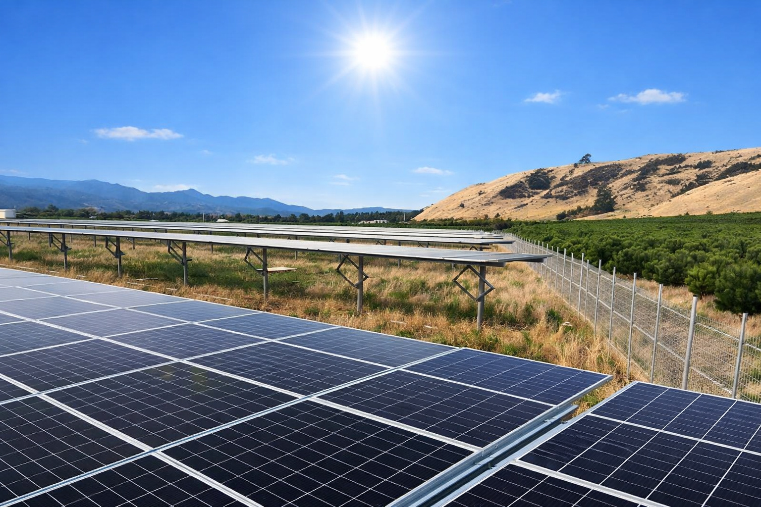 Usina de energia solar com fileiras de painéis sob sol forte e céu azul ao meio-dia, com montanhas ao fundo.