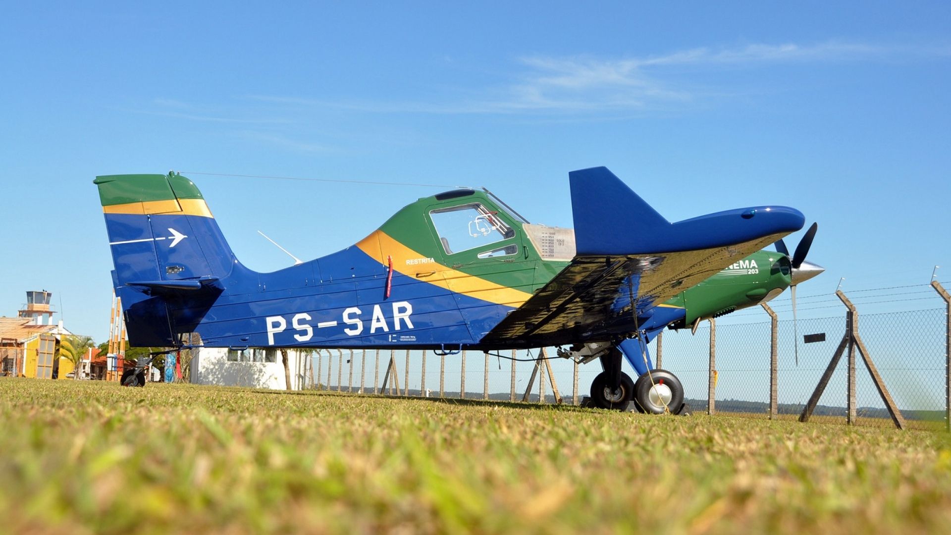 Avião agrícola Ipanema da Embraer, pintado nas cores do Brasil, estacionado em pista gramada sob céu azul, utilizado em operações do agronegócio com foco em sustentabilidade.