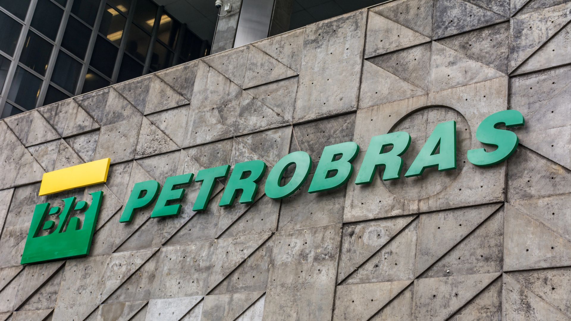 Logotipo da Petrobras em destaque na fachada de um edifício corporativo, com letras verdes e símbolo da empresa aplicados sobre parede de concreto.