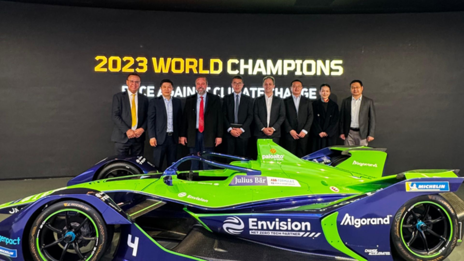 Executivos em evento da Envision posam ao lado de carro da Fórmula E, símbolo de tecnologia limpa e inovação em energia sustentável