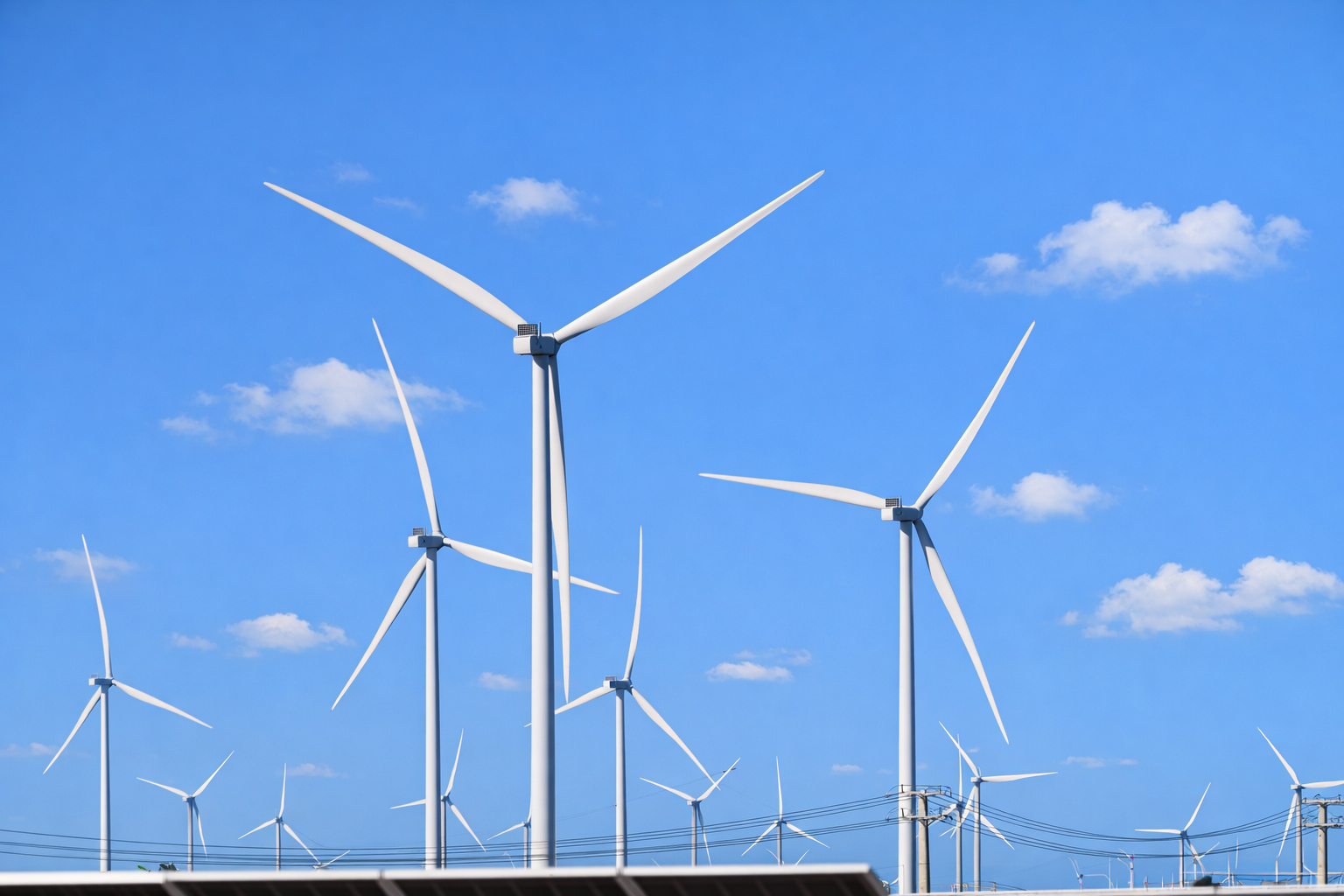 Parque eólico com vários aerogeradores brancos sob céu azul com poucas nuvens, visto a partir do nível do solo, com linhas de transmissão ao fundo.
