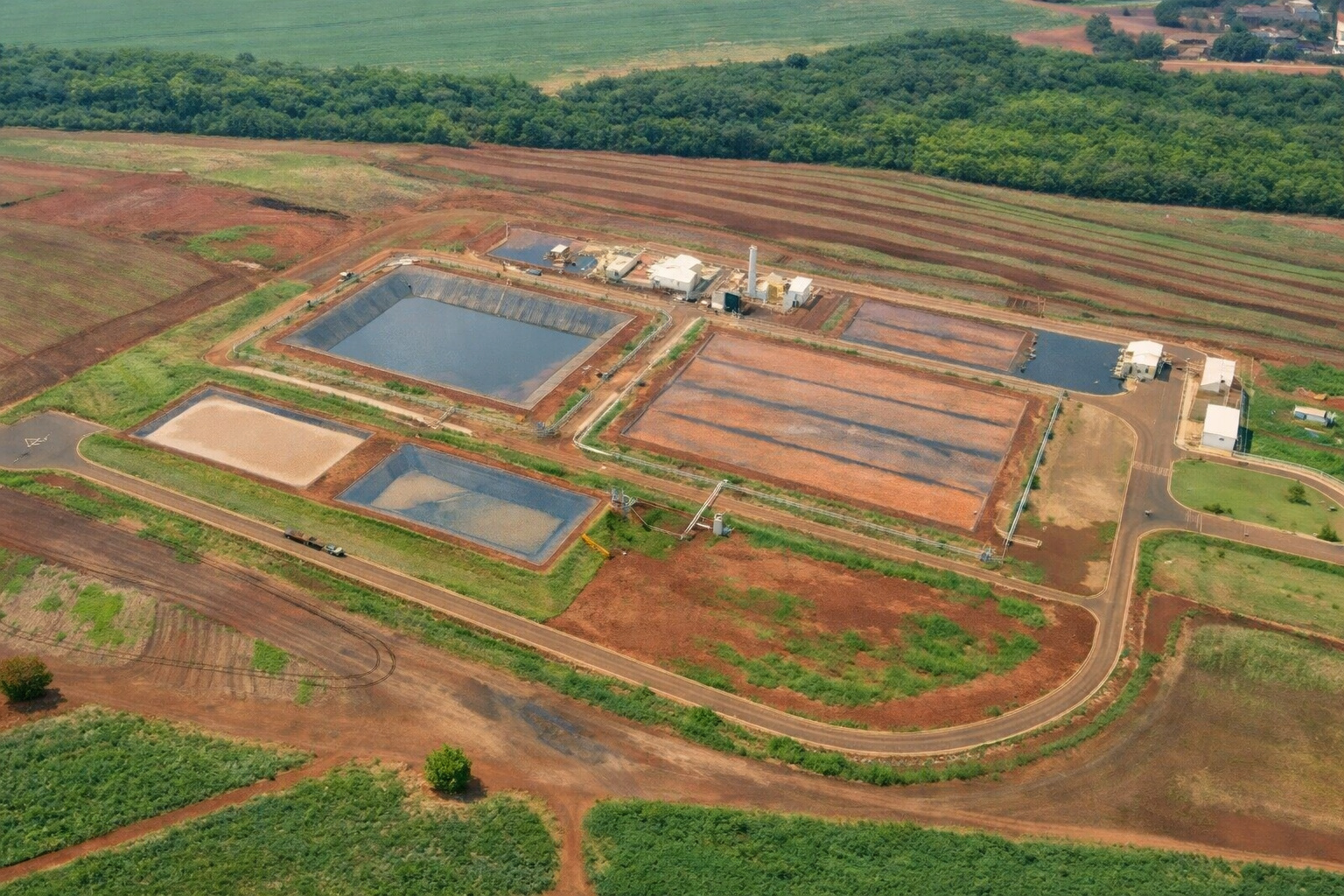Vista aérea de estação de tratamento com tanques retangulares, estradas internas e áreas agrícolas ao redor sob luz forte da tarde.