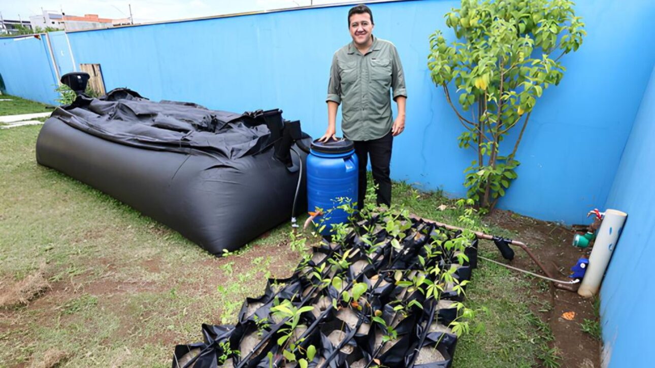 Hombre Al Lado De Un Sistema De Biogás Compuesto Por Un Barril Azul, Estructura Inflable Negra Para Almacenamiento De Gas Y Huerta Comunitaria En Recipientes De Tela, Instalada En Escuela De São José Dos Campos.