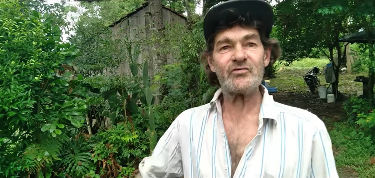 Homem vivendo isolado em área rural aparece em frente a rancho simples cercado por vegetação, sem acesso à energia elétrica
