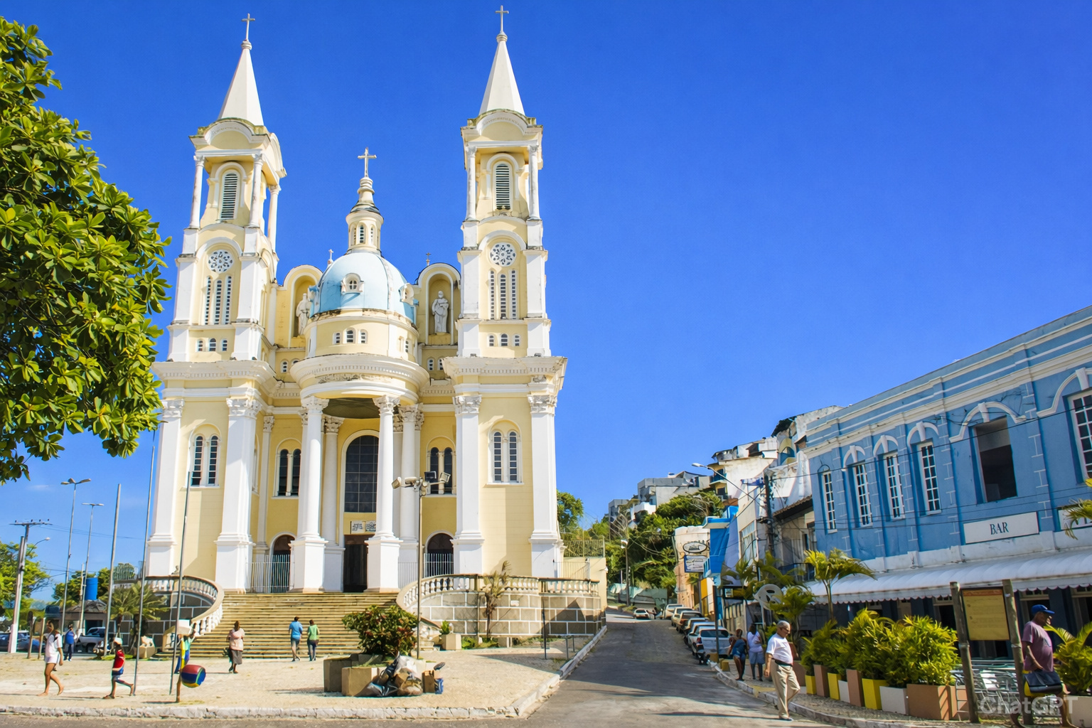 Igreja católica de fachada amarela e branca com duas torres e cúpula central sob céu azul sem nuvens, em área urbana com pessoas caminhando.