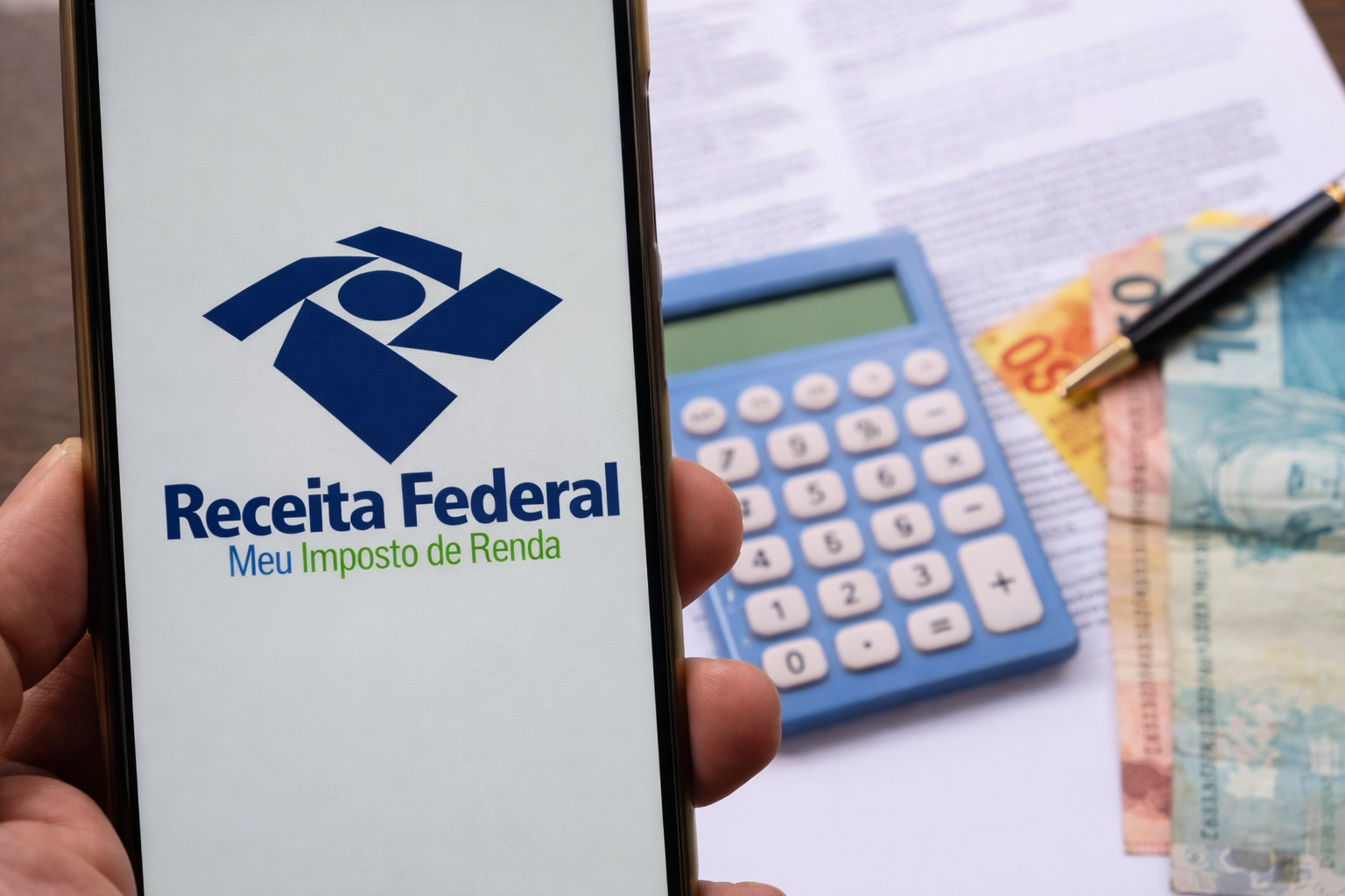 Pessoa segurando um smartphone com o aplicativo da Receita Federal aberto, com calculadora e documentos ao fundo.