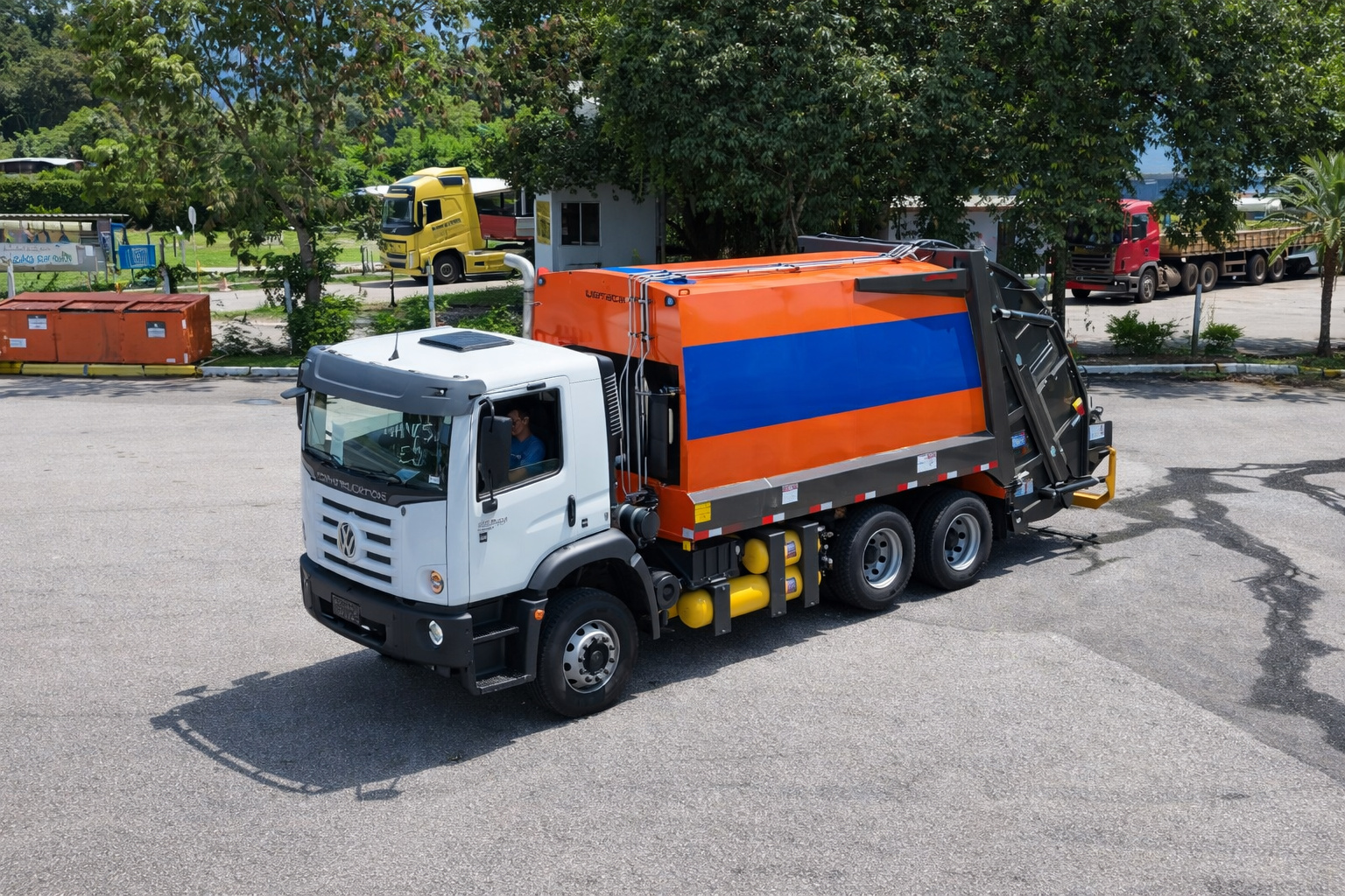 Caminhão de coleta de lixo branco com compartimento laranja e azul estacionado em pátio ao ar livre sob iluminação de meio-dia.